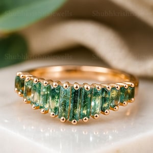 May include: A gold ring with a row of rectangular, green gemstones. The ring has a delicate design with small, gold accents. The gemstones are a vibrant green colour, set in a gold band. The ring is displayed on a white surface.