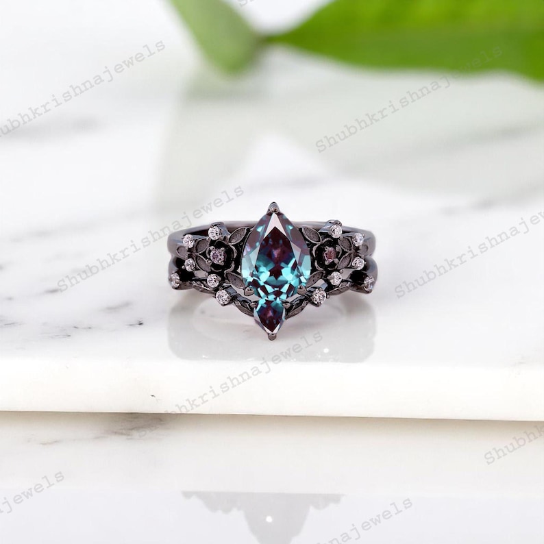May include: A black metal ring set with a marquise-cut, teal-colored gemstone. The ring is adorned with floral and small diamond accents. The band has a matching floral design. The ring is set on a white surface.