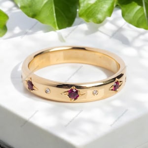 Może przedstawiać: Złoty pierścionek z obrączką z naprzemiennymi, gwiaździstymi oprawami z okrągłymi, głęboko czerwonymi kamieniami szlachetnymi i małymi, okrągłymi, przezroczystymi kamieniami szlachetnymi. Pierścionek jest osadzony na białej powierzchni z zielonymi liśćmi w tle.