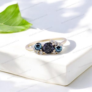 Könnte beinhalten: Ein goldener Ring mit drei blauen Edelsteinen. Der mittlere Stein ist ein großer, runder, dunkelblauer Stein mit einer funkelnden, strukturierten Oberfläche. Die beiden kleineren Steine sind rund und in einem helleren Blauton. Die Steine sind in einer goldenen Fassung mit kleinen, weißen Diamanten eingefasst.