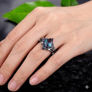 May include: A dark, ornate ring featuring a large, square-cut, color-changing gemstone with blue and purple hues. The ring has a black metal band with floral and leaf detailing, and small white accent stones.