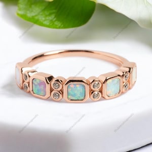 May include: A rose gold band ring with five square-shaped opal gemstones and small diamond accents. The ring is set on a white background.