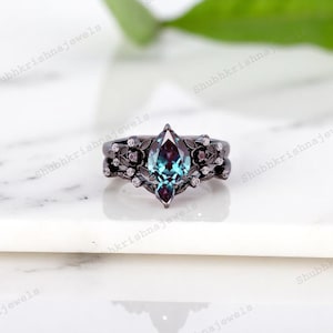 May include: A black metal ring set with a marquise-cut, teal-colored gemstone. The ring is adorned with floral and small diamond accents. The band has a matching floral design. The ring is set on a white surface.