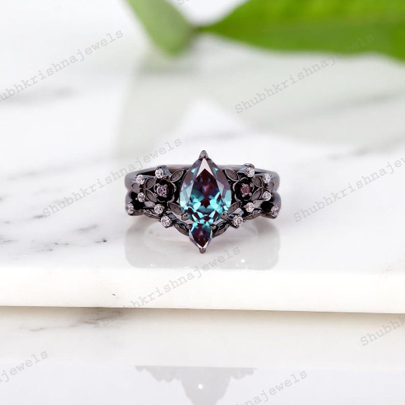 May include: A black metal ring set with a marquise-cut, teal-colored gemstone. The ring is adorned with floral and small diamond accents. The band has a matching floral design. The ring is set on a white surface.