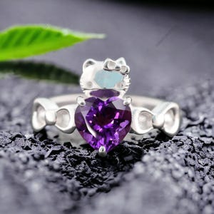 May include: A silver ring with a purple heart-shaped gemstone and a small white cat charm. The ring is set with a Celtic knot design.