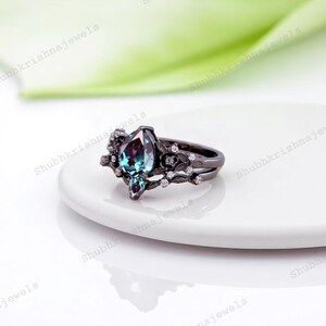 May include: A black ring with a large, pear-shaped, teal and purple gemstone as the centerpiece. The ring features a floral design with small white gemstones and is set on a white surface.