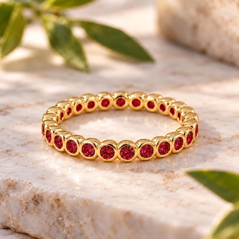 May include: A gold eternity band ring featuring a continuous row of round, bezel-set red gemstones. The metal has a polished finish, and the stones are uniform in size and colour, creating a circular design that encircles the entire band.