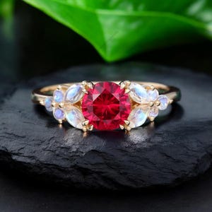May include: A gold ring with a large, round, red gemstone at its centre. The ring is decorated with smaller, petal-shaped, white and blue gemstones on either side of the main stone. The ring is displayed against a dark background.