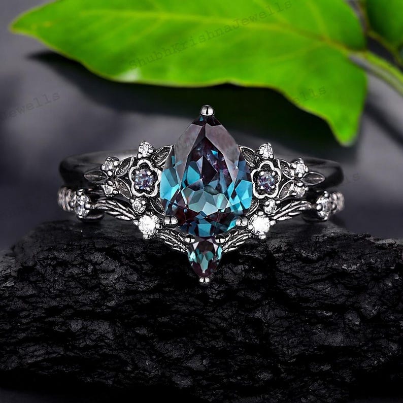 May include: A two-piece ring set featuring a pear-shaped, color-changing alexandrite gemstone. The ring is set in a dark metal with floral and leaf accents, and small white gemstones. The band has a matching design.