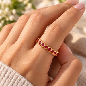 May include: A gold band ring featuring a continuous row of round, vibrant red stones set in individual circular bezels. The ring has a polished metallic finish and a uniform design that encircles the finger with consistent spacing.
