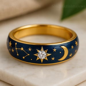 Op de afbeelding: Een goudkleurige ring met een donkerblauwe emaille band met hemelse ontwerpen. De ring heeft een centraal sterrenpatroon met een heldere edelsteen, een maan en diverse sterren. De binnenband van de ring is ook goudkleurig.