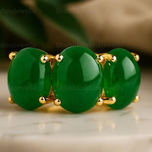 Puede incluir: Un anillo de oro con tres grandes piedras preciosas ovaladas de color verde vibrante. El anillo tiene pequeños detalles dorados y se exhibe sobre una superficie de mármol blanco. Las piedras están engastadas en una banda de oro.