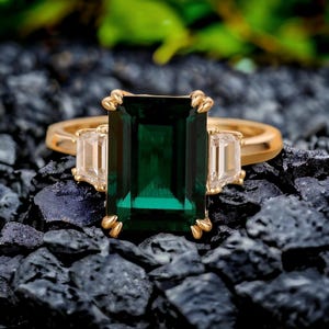 May include: A gold ring featuring a large emerald green gemstone flanked by two smaller clear gemstones.