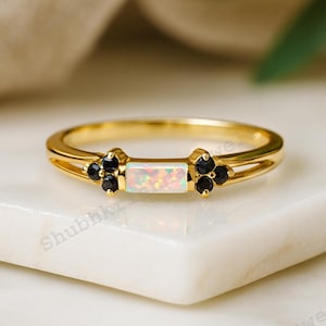 Può includere: Un anello d'oro con una pietra centrale di opale rettangolare e accenti di pietre nere. L'anello ha un design a gambo diviso ed è posto su una superficie bianca. L'opale mostra un gioco di colori. Un gioiello.