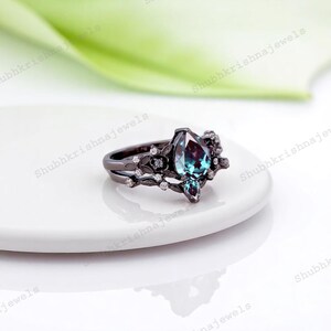 May include: A black ring with a pear-shaped, color-changing gemstone. The ring features a floral design with small, clear accent stones. The band is split, and the ring sits on a white surface.