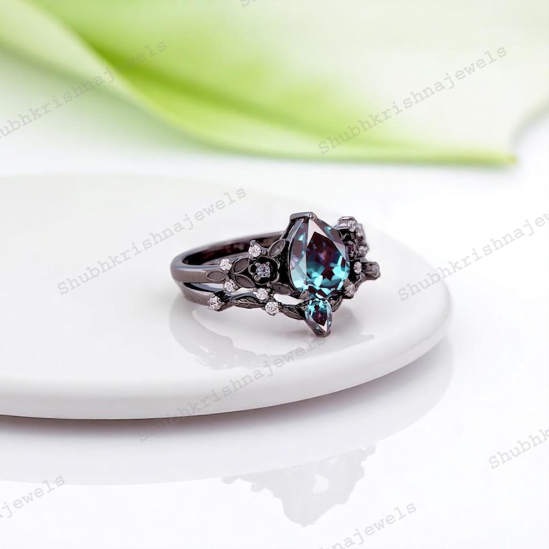 May include: A black ring with a pear-shaped, color-changing gemstone. The ring features a floral design with small, clear accent stones. The band is split, and the ring sits on a white surface.