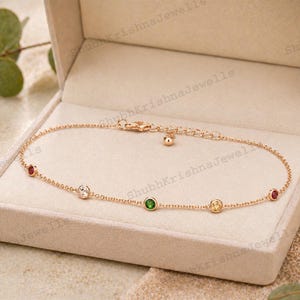 Puede incluir: Una delicada tobillera dorada con una serie de gemas de colores, incluyendo piedras rojas, blancas, verdes y amarillas, se muestra dentro de una caja de joyería beige. La tobillera está finamente elaborada con un pequeño dije cerca del cierre.