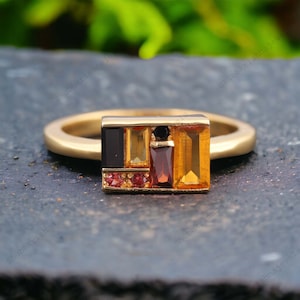 Puede incluir: Un anillo de oro con un engaste rectangular que presenta una variedad de piedras preciosas de colores, incluyendo negro, rojo y naranja.