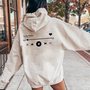 May include: A white hoodie with a black music player graphic on the back. The graphic includes a heart, play button, and rewind and fast forward buttons. The text "Song Name" and "Artist Name" are also included in the graphic.