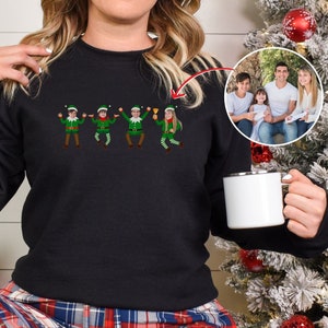 Op de afbeelding: Zwart sweatshirt met een afbeelding van vijf dansende elven. De elven dragen groene hoeden, groene shirts en rode broeken. Het sweatshirt heeft een ronde hals en lange mouwen.