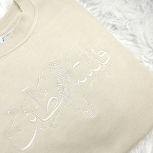 May include: A beige sweatshirt with white embroidery of Arabic calligraphy. The calligraphy reads "فَلَسْتَ عَلَيْنَا بِشَيْءٍ".