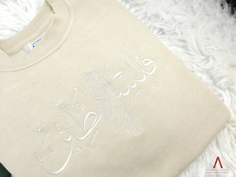 May include: A beige sweatshirt with white embroidery of Arabic calligraphy. The calligraphy reads "فَلَسْتَ عَلَيْنَا بِشَيْءٍ".