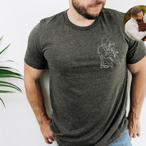 以下が含まれることがあります： 子供を抱いている人の白い線画が特徴の濃い灰色のTシャツ。右上隅にある小さな円形の画像は、子供を抱いている人を示しています。シャツは柔らかい素材で作られています。