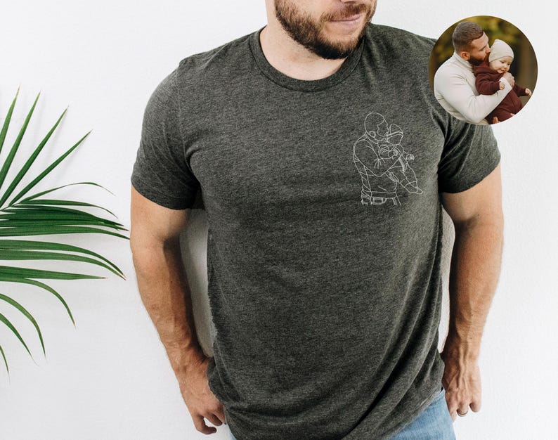 以下が含まれることがあります： 子供を抱いている人の白い線画が特徴の濃い灰色のTシャツ。右上隅にある小さな円形の画像は、子供を抱いている人を示しています。シャツは柔らかい素材で作られています。