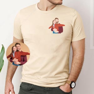 Pu&ograve; includere: Una maglietta beige chiaro con una piccola grafica di una coppia che si abbraccia sulla parte superiore sinistra del petto. Un'immagine circolare pi&ugrave; grande della coppia &egrave; visibile sotto. La persona che indossa la maglietta indossa anche un orologio.