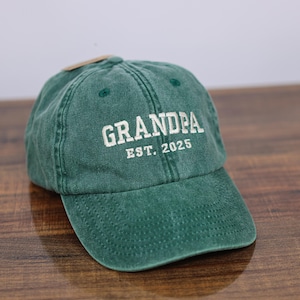 May include: A green cotton baseball cap with white embroidered text that reads "GRANDPA EST. 2025".