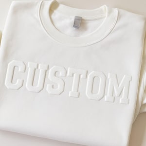 Op de afbeelding: Witte trui met het woord "CUSTOM" in reliëf.