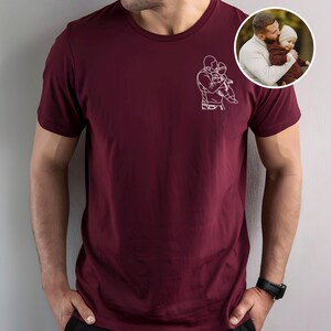 以下が含まれることがあります： 親が子供を抱いている白い線画が特徴のマルーンTシャツ。右上隅にある小さな円形の画像には、親と子供が写っています。シャツは半袖で、クルーネックです。