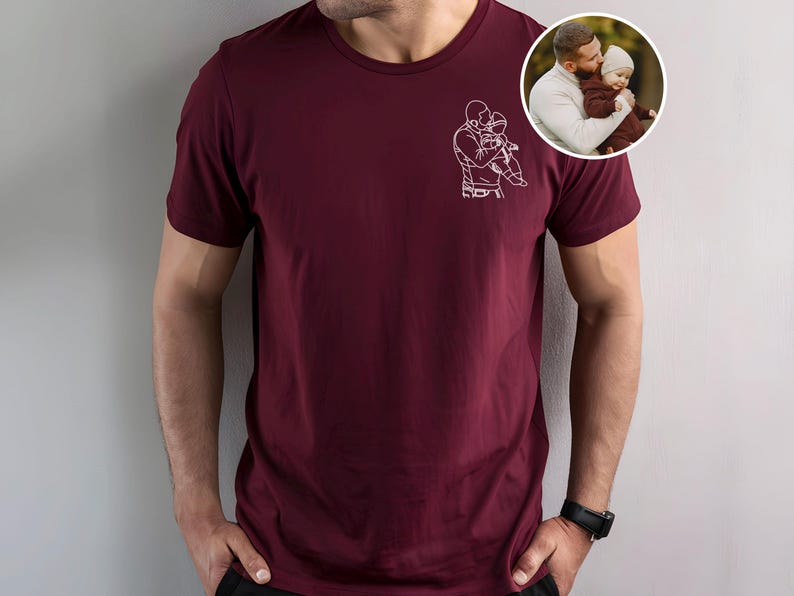 以下が含まれることがあります： 親が子供を抱いている白い線画が特徴のマルーンTシャツ。右上隅にある小さな円形の画像には、親と子供が写っています。シャツは半袖で、クルーネックです。