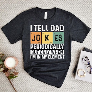 May include: Dark gray t-shirt with the text "I TELL DAD JOKES PERIODICALLY BUT ONLY WHEN I'M IN MY ELEMENT" in white. The word "JOKES" is in a periodic table style with orange, yellow, and green blocks.