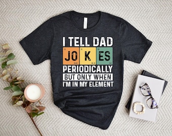 Puede incluir: Camiseta gris oscuro con el texto "I TELL DAD JOKES PERIODICALLY BUT ONLY WHEN I'M IN MY ELEMENT" en blanco. La palabra "JOKES" está en un estilo de tabla periódica con bloques de colores.