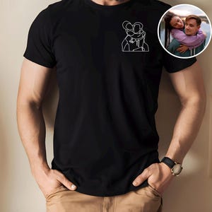 以下が含まれることがあります： 2 人が抱き合っている白い線画が特徴の黒い T シャツ。右側には、カップルの小さな円形の画像が見えます。画像内の人物は黒い時計とカーキ色のズボンを着用しています。