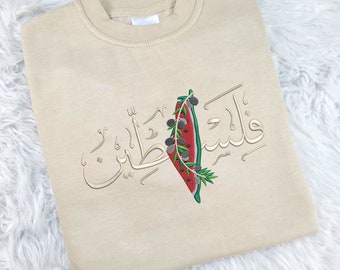 May include: A beige sweatshirt with an embroidered design featuring a watermelon, olive branches, and Arabic text that reads "Palestine".