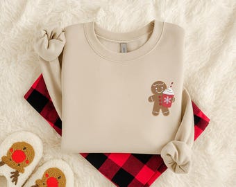 Pode incluir: Camisola bege com um desenho de homem de gengibre segurando uma xícara de café com um floco de neve. A camisola é combinada com calças de pijama xadrez vermelho e preto e pantufas de rena.