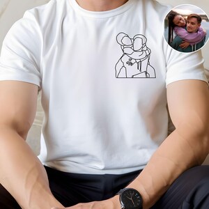 以下が含まれることがあります： 2 人が抱き合っているミニマリストの線画イラストが特徴の白いTシャツ。小さな円形の画像には、抱き合っているカップルが写っています。シャツは柔らかく明るい色の生地で作られています。その人は黒い腕時計をしています。