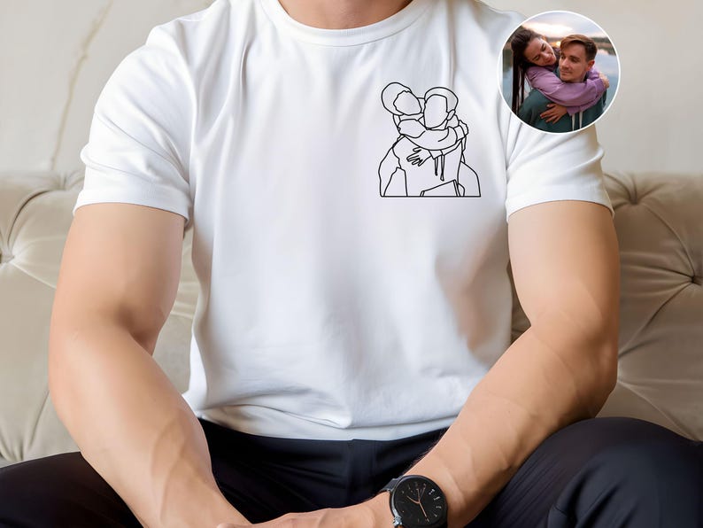 以下が含まれることがあります： 2 人が抱き合っているミニマリストの線画イラストが特徴の白いTシャツ。小さな円形の画像には、抱き合っているカップルが写っています。シャツは柔らかく明るい色の生地で作られています。その人は黒い腕時計をしています。