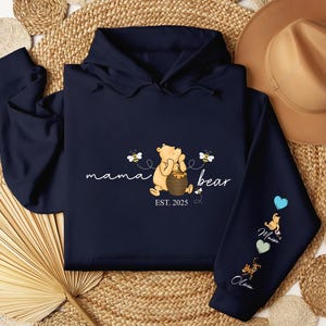 May include: A navy blue hoodie with a Winnie the Pooh illustration and the text "mama bear est. 2025". The hoodie has three different colored patches on the sleeve with the names "Olivia", "Maxine", and "Charlie" on them.