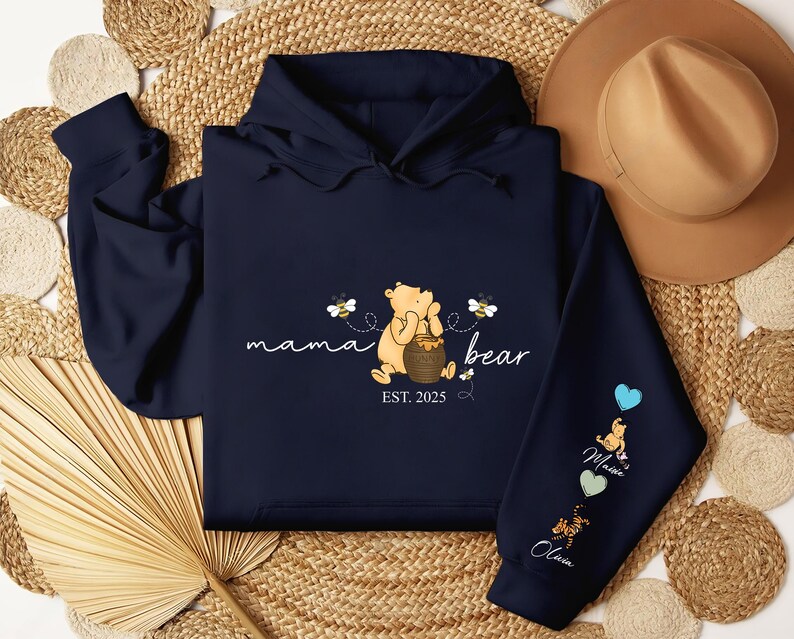 May include: A navy blue hoodie with a Winnie the Pooh illustration and the text "mama bear est. 2025". The hoodie has three different colored patches on the sleeve with the names "Olivia", "Maxine", and "Charlie" on them.