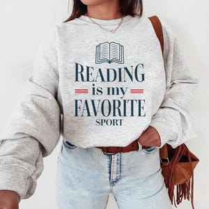 May include: A gray sweatshirt with the text "Reading is my favorite sport" and an illustration of an open book. The text is in navy blue with red lines under "is my".