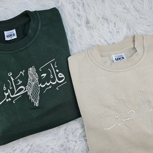 May include: Two sweatshirts with Arabic calligraphy and a stylized image of Palestine. The sweatshirt on the left is dark green and the sweatshirt on the right is beige.