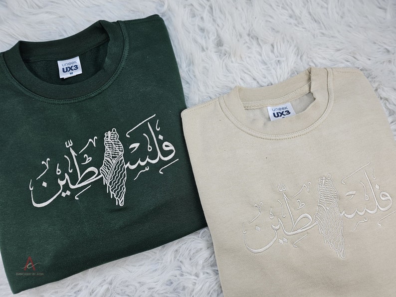 May include: Two sweatshirts with Arabic calligraphy and a stylized image of Palestine. The sweatshirt on the left is dark green and the sweatshirt on the right is beige.