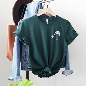 Pu&ograve; includere: T-shirt verde scuro con una piccola illustrazione di un genitore e un bambino che si tengono per mano. La maglietta &egrave; esposta su un appendiabiti con una giacca di jeans, una borsa di pelle marrone e un cappello sullo sfondo. La maglietta ha le maniche arrotolate.