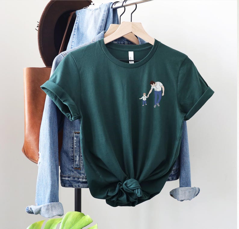 Pu&ograve; includere: T-shirt verde scuro con una piccola illustrazione di un genitore e un bambino che si tengono per mano. La maglietta &egrave; esposta su un appendiabiti con una giacca di jeans, una borsa di pelle marrone e un cappello sullo sfondo. La maglietta ha le maniche arrotolate.