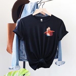 Pu&ograve; includere: T-shirt nero con una piccola grafica di una coppia che si abbraccia. La maglietta &egrave; appesa a un attaccapanni con una giacca di jeans, una borsa di pelle marrone e un cappello marrone. La maglietta ha maniche corte e un orlo annodato.