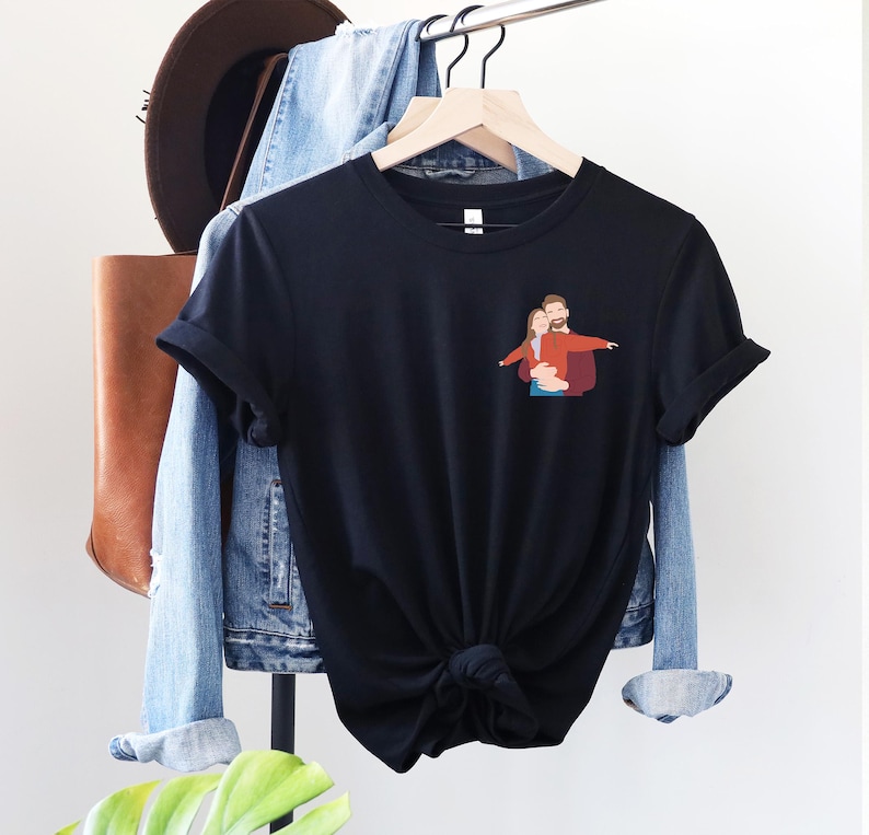 Pu&ograve; includere: T-shirt nero con una piccola grafica di una coppia che si abbraccia. La maglietta &egrave; appesa a un attaccapanni con una giacca di jeans, una borsa di pelle marrone e un cappello marrone. La maglietta ha maniche corte e un orlo annodato.