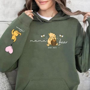 May include: Green hooded sweatshirt with a Winnie the Pooh inspired design. The text reads "mama bear est. 2023" with a bear holding a honey pot and bees flying around. The left sleeve has a Winnie the Pooh character holding a pink heart balloon.